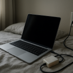Laptop charger resting on a bed beside an open laptop